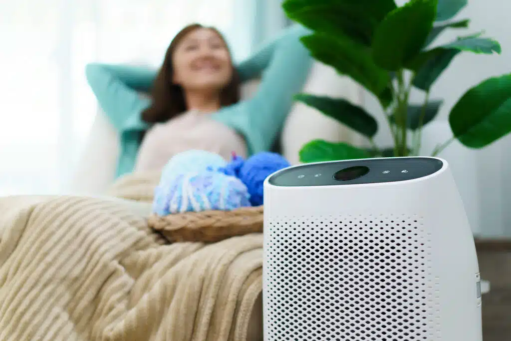 A happy homeowner relaxing on a couch in the background, with an air purifier in the foreground